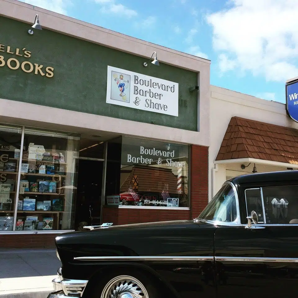 Exterior of Boulevard Barber & Shave storefront in a shopping plaza. A large sign reading “Boulevard Barber & Shave” is mounted above the entrance, with the shop name also displayed on the front window. A vintage black car is parked in the foreground, partially blocking the lower portion of the building. A bookstore window is visible next door, and the scene is lit by daylight under a partly cloudy sky.