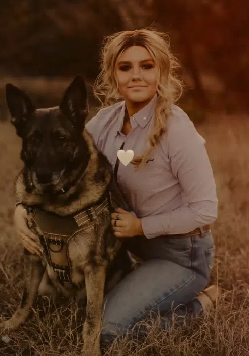 Wigs For Kids recipient Gauge kneels in a field of tall, dry grass beside a large German Shepherd wearing a harness. She has long, light-colored hair styled in a loose side braid and is dressed in a light button-down shirt and jeans.