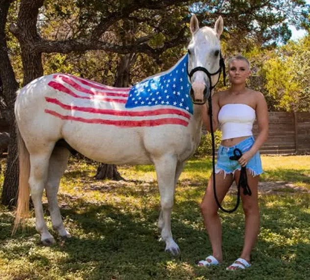 A white horse stands outdoors beneath large trees with an American flag painted across its side. Gauge, with head-shaved and wearing a white strapless top, denim shorts, and white sandals stands beside the horse holding its lead rope. Sunlight filters through the trees, illuminating the grassy area and wooded background.