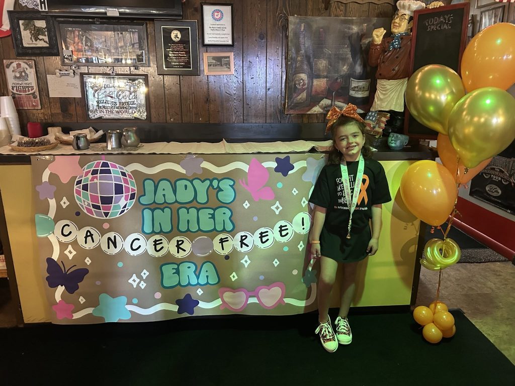 Wigs For Kids Recipient Jady poses at her bell-ringing celebration. The banner nehind her reads "Jady's in her Cancer-Free Era."