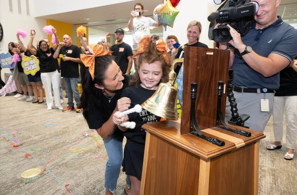 7 year old Jady rings a ceremonial bell at a hospital while supporters, family members, and a television news crew gather to celebrate the completion of cancer treatment.