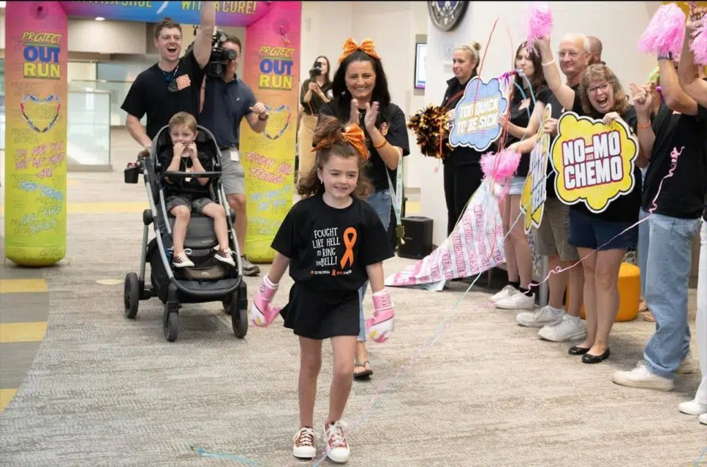 A young child, 7 year old Jady, is wearing pink boxing gloves walks through a hospital hallway lined with supporters holding signs and pom-poms during a celebration marking the completion of cancer treatment.