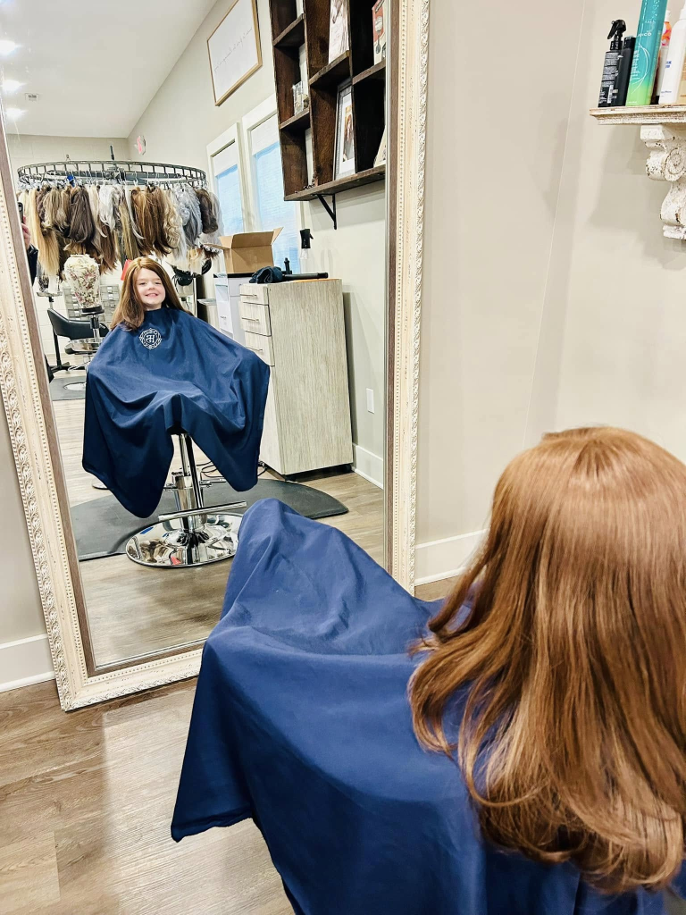 Jady, a Wigs For Kids recipient, sits in a salon chair wearing a styling cape and looking into a large floor mirror. The image is photographed from behind, showing Jady's newly fitted long brown hair replacement system while her reflection is visible in the mirror. In the background, a salon workstation and a display of hairpieces can be seen. The photo captures Jady seeing her appearance with her new hair replacement system during a Wigs For Kids salon appointment.