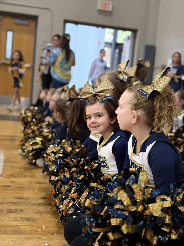 Wigs For Kids Recipient Jady smiles toward the camera, with her natural hair regrowing and tied with ribbon. She is dressed in blue and gold cheerleading outfit. She is seated in a line with other cheerleaders in a school gymnasium.