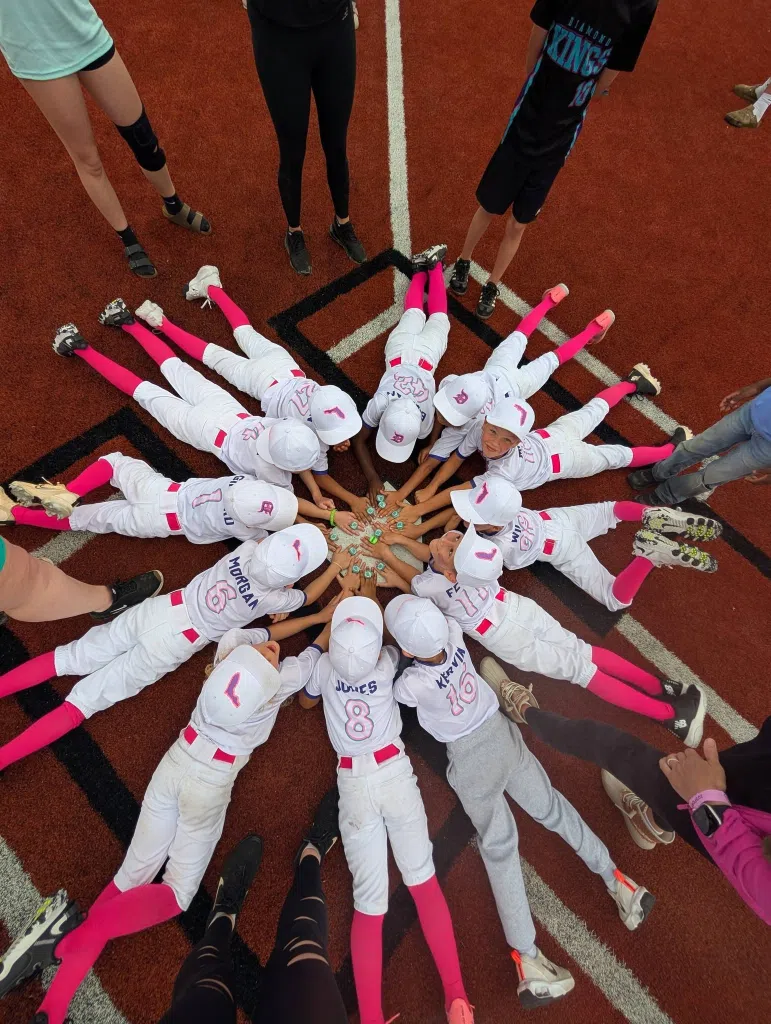 Members of a youth baseball team lie in a circle on the field with their hands joined in the center, displaying championship and MVP rings after a tournament victory.
