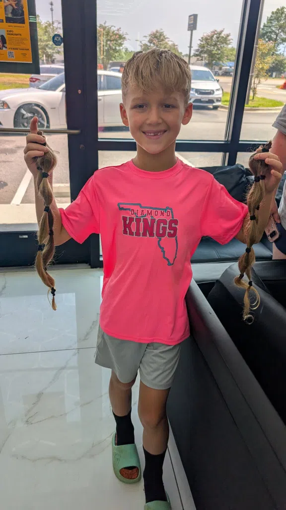 7 year old Wigs For Kids hair donor Pryor stands proudly holding his blonde ponytails, freshly cut for donation.