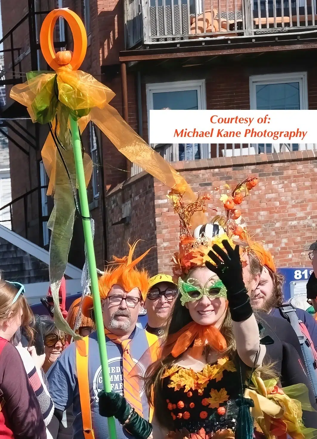 Pumpkin Queen Sarah Wentworth wearing an elaborate autumn-themed costume and decorative green mask walks in a community parade while carrying a tall staff topped with a pumpkin and flowing orange and green ribbons. The costume includes fall leaves, seasonal decorations, and long gloves. Several parade attendees and spectators are visible behind the participant, with brick buildings lining the street. A photo credit reading “Courtesy of: Michael Kane Photography” appears in the upper right corner.