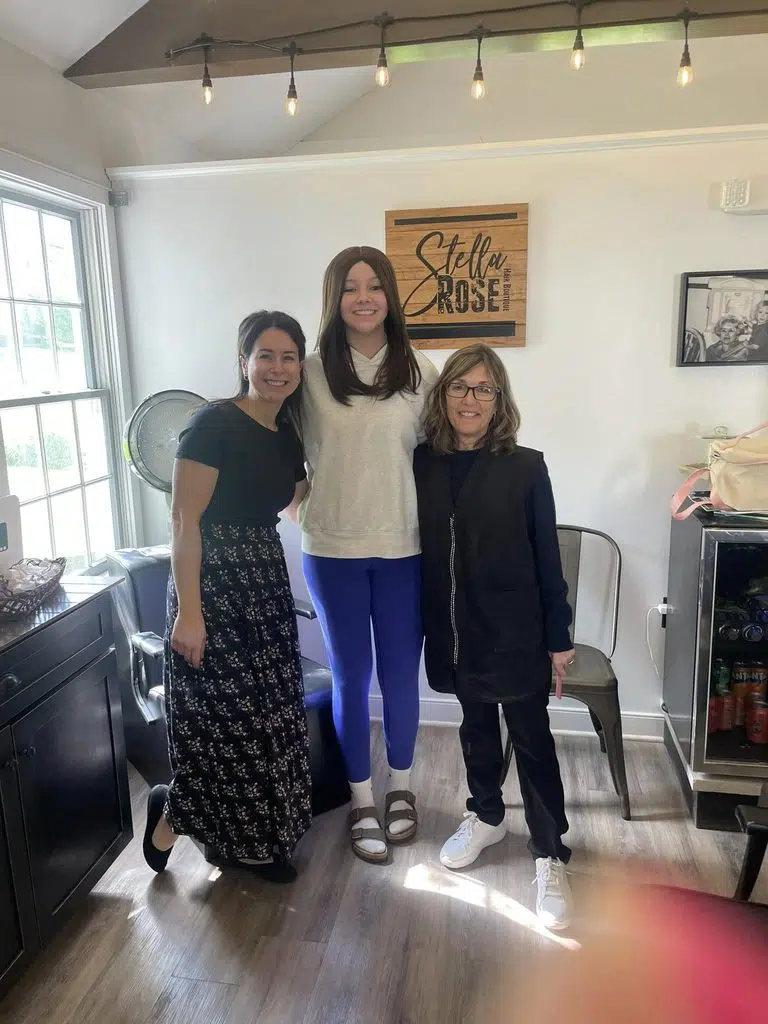 Wigs For Kids free hairpiece recipient Gabby stands between her two volunteer stylists, Tori Wiltrout and Michelle Licovoli-Preston of Stella Rose boutique. Gabby is wearing a brown hair wig gifted through the Wigs For Kids Recipient program, smiling.