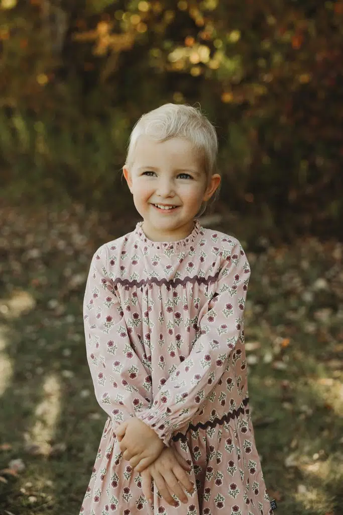 Photo of 4 -year-old Cece. She is wearing her natural hair, which is a very short light blonde. She is wearing a patterned dress and is smiling in a nature setting.