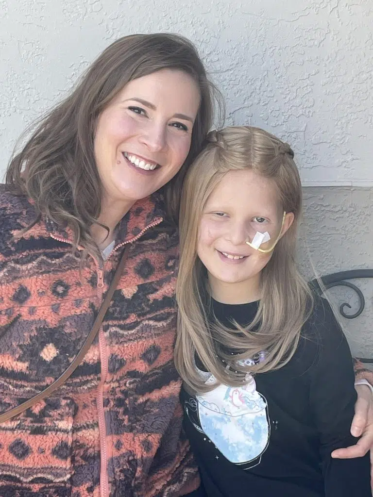 Ellie, the subject of this Wigs For Kids story, sits beside her mother for a portrait outdoors. Ellie wears a black shirt and has a feeding tube visible on her cheek, reflecting part of her childhood cancer treatment journey. Her mother has an arm around her as they sit together against a light-colored wall. The image highlights the close support of family throughout Ellie's battle with a rare brain tumor and her path toward recovery.