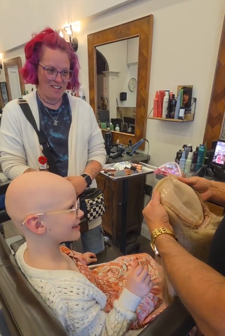 Wigs For Kids recipient Aurora is seated in a stylist's chair, smiling. She is bald due to alopecia, and is looking at a blonde wig that was made for her at no charge from Wigs For Kids. Her mother looks on in approval in the background.