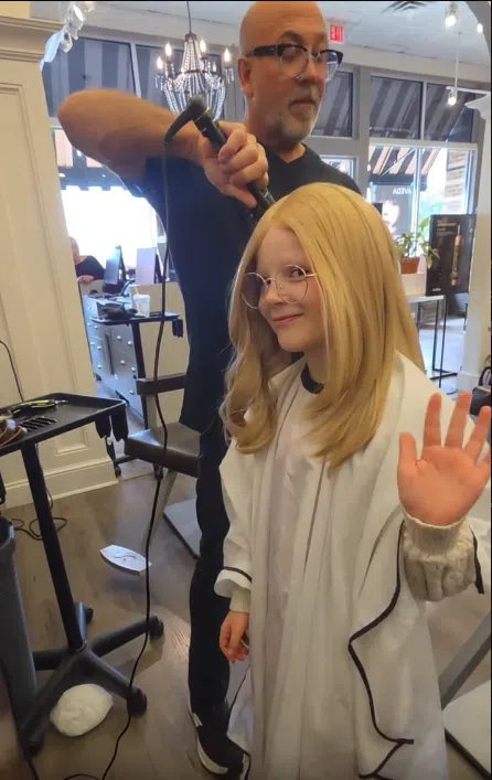 Wigs For Kids recipient Aurora is standing in a hair salon, wearing her blonde hair wig provided at no cost to her family. Her stylist George stands behind her, curling sections of the wig. Aurora is smiling and waving at the camera.