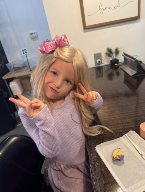 4-year-old Cece is posing for the camera, her long blonde wig styled with a bow in her hair. She is holding 2 fingers up in a peace sign and looks happy.