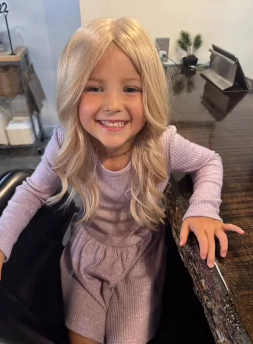 Cece is wearing her long blonde Wigs For Kids hairpiece in a salon chair and is smiling at the camera. It is lightly curled and lays past her shoulders.